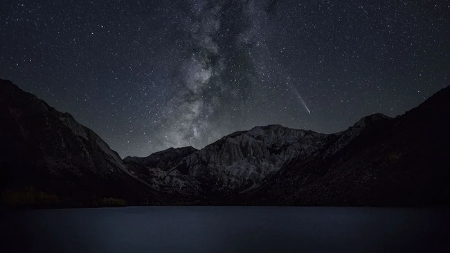 Milky Way galaxy arches over a dark mountain landscape reflected in still water below.