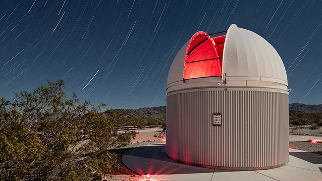 Observatory dome with open slot revealing illuminated red telescope against star trail sky.