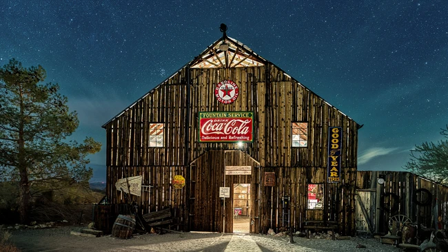 Weathered wooden general store at night beneath a starlit sky, illuminated by warm interior light and vintage neon signage.