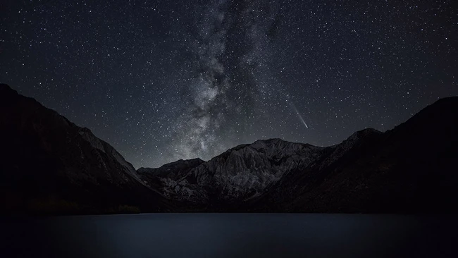 Milky Way galaxy stretches across a starlit night sky above snow-capped mountains and a dark alpine lake.