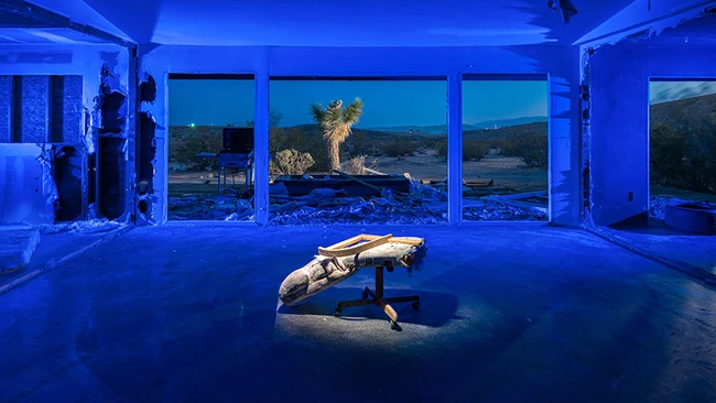 Person in plank position on bench inside blue-lit studio with large windows overlooking desert landscape.