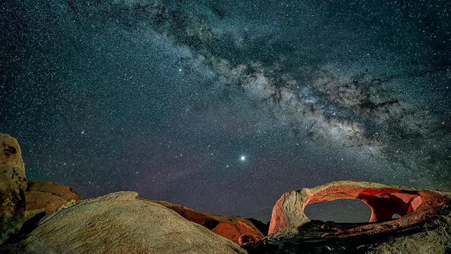 Milky Way galaxy stretching across night sky above desert rock formations with natural arch.