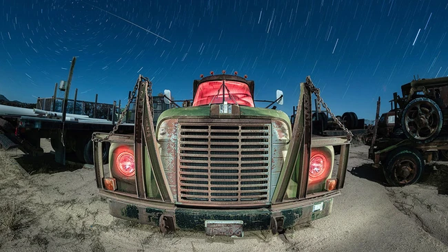 Abandoned truck with red cab and green hood photographed head-on at night with star trails streaking across the sky.