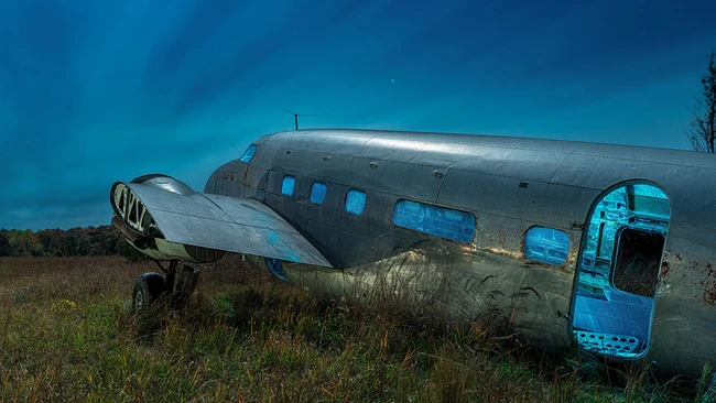 Abandoned aircraft fuselage with detached wing panel sits in overgrown field under dramatic blue sky.