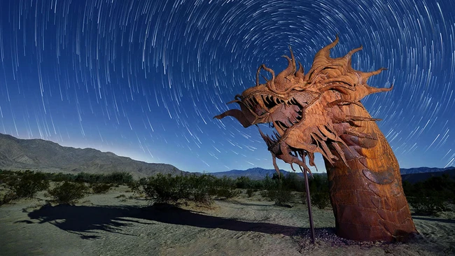 Large wooden dragon sculpture in desert landscape beneath circular star trails from long exposure.