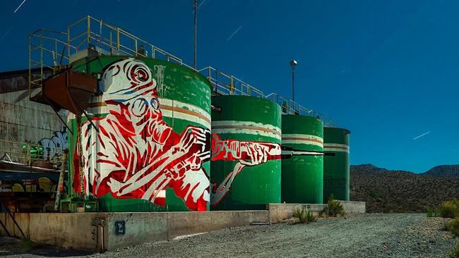 Large industrial storage tanks with bold red and white street art mural against clear blue sky.