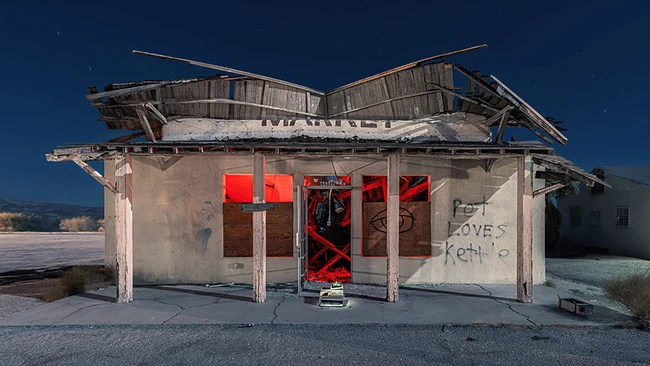 Abandoned roadside diner with boarded red doors and deteriorating wooden roof against clear blue sky.