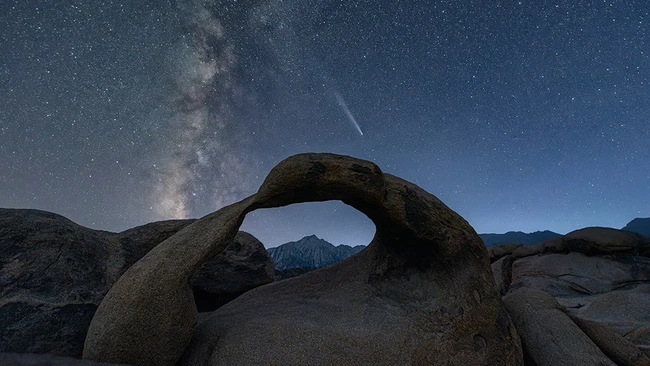 Massive boulder with natural arch framing distant mountains under starry night sky with Milky Way visible.