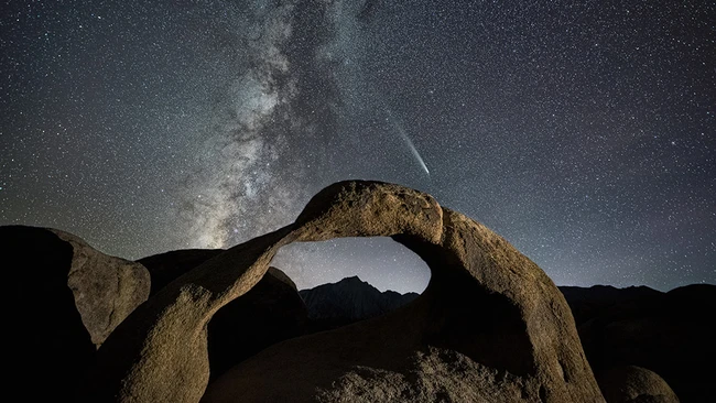 Milky Way galaxy arching over a natural rock arch formation under a starry night sky with a visible meteor.