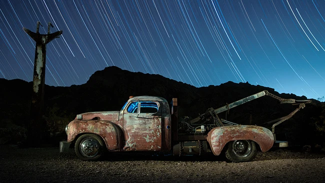 Two rusted vintage pickup trucks positioned side-by-side in a desert landscape at night, with star trails streaking across the dark sky and a cross silhouetted on a distant hill.