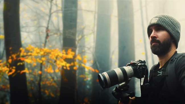 Bearded photographer in knit beanie holding telephoto lens in misty forest with golden foliage