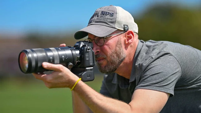 Photographer with white cap holding DSLR camera with telephoto lens, shooting outdoors in bright sunlight