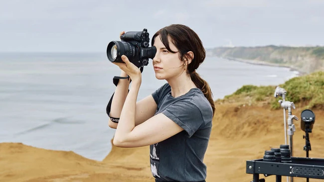 Female photographer holding DSLR camera to her face while standing on coastal cliff overlooking ocean