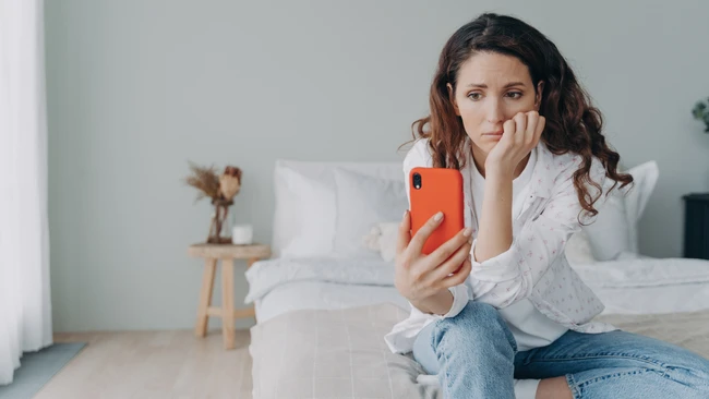 Woman in white shirt and jeans sitting on bed holding orange smartphone