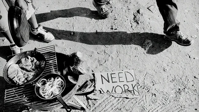 Overhead shot of person sitting on pavement with food and sign reading "NEED WORK"
