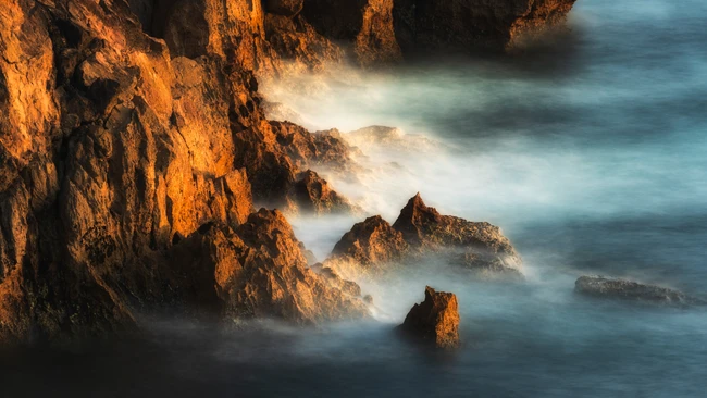 Golden-lit coastal cliffs with misty waves and rocky outcrops at golden hour