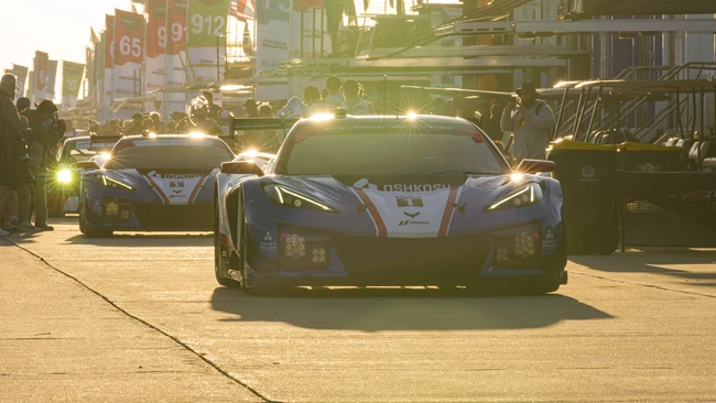 White and blue sports car leading a parade of racing vehicles down an urban street in golden hour sunlight