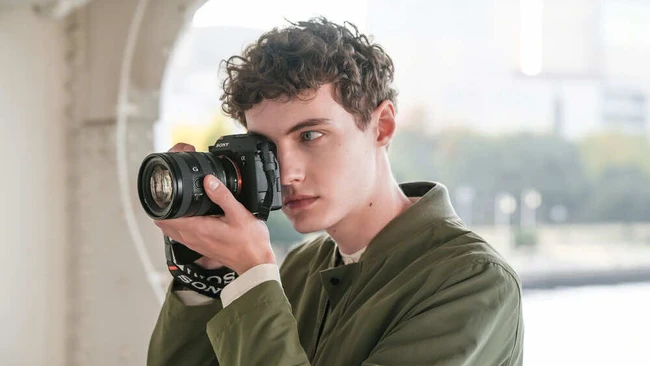 Young photographer holding DSLR camera with telephoto lens to eye while looking toward camera