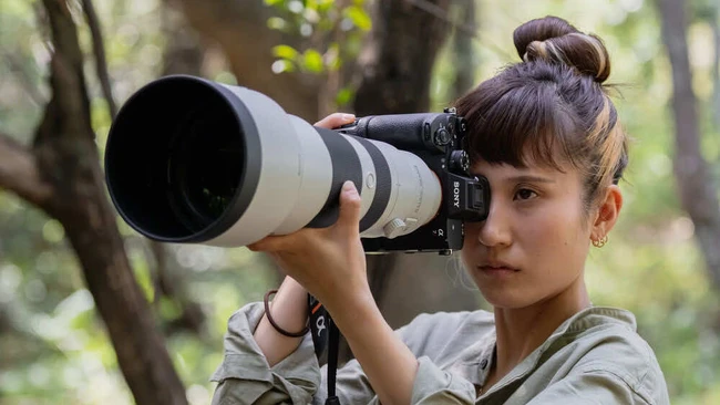 Young woman holding a telephoto lens to her eye in a wooded outdoor setting