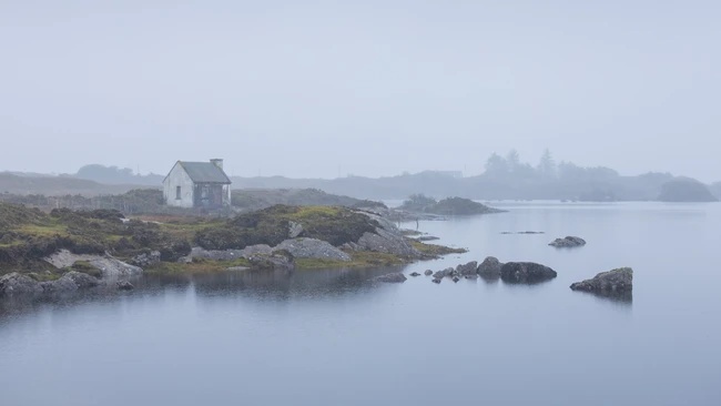 Isolated white house on rocky coastal peninsula shrouded in morning mist