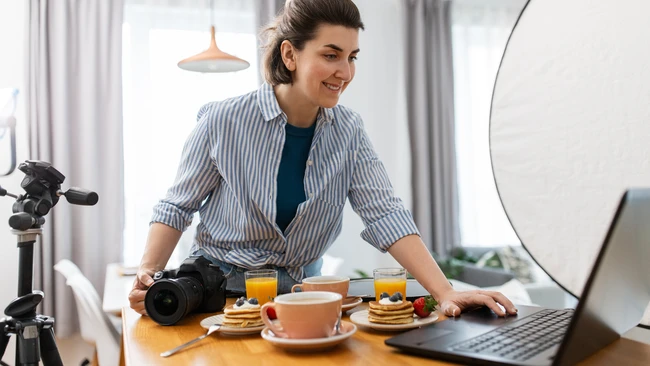 Female content creator at wooden desk with professional DSLR camera, ring light, and laptop during food photography setup