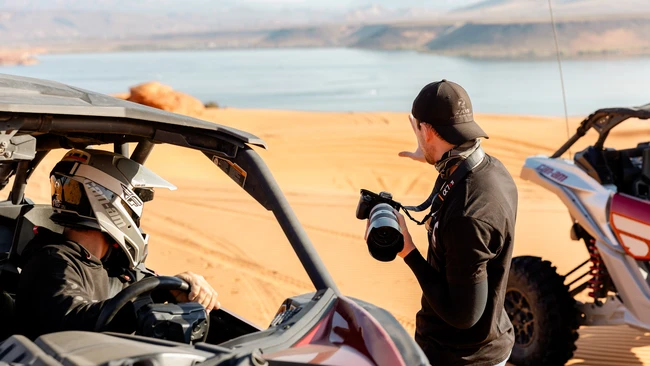 Photographer with telephoto lens shooting beside off-road vehicles overlooking desert reservoir landscape