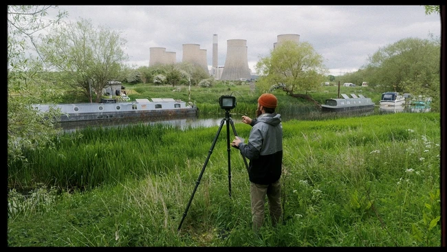 Photographer operating a surveying theodolite on tripod in green field with industrial cooling towers in background
