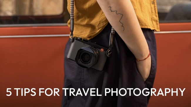 Photographer wearing yellow shirt and dark pants holding compact camera with chain strap against rust-colored wall