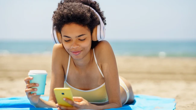 Woman wearing headphones lying on beach towel holding smartphone and beverage