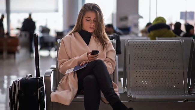 Woman in beige cardigan seated in modern airport terminal holding smartphone