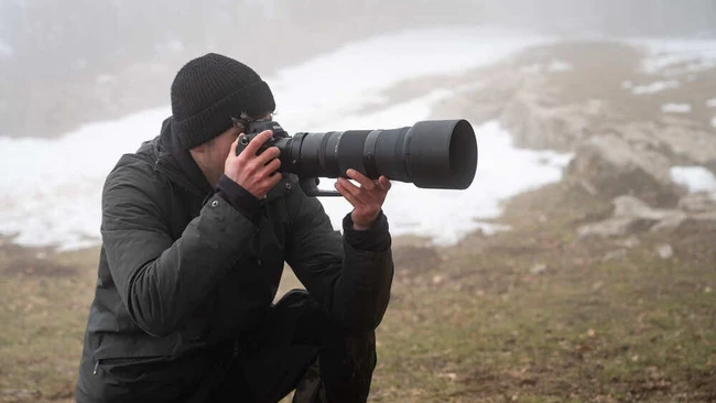 Photographer in black winter gear crouching while composing shot with telephoto lens in snowy mountain terrain