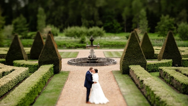 Bride and groom kissing on a formal garden pathway lined with manicured hedges and topiary cones