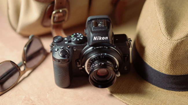 Nikon rangefinder camera with external viewfinder and lens, surrounded by sunglasses and straw hat on neutral fabric backdrop