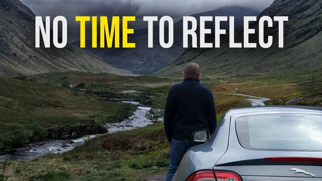 Person standing beside a silver car overlooking a mountain valley with a winding stream