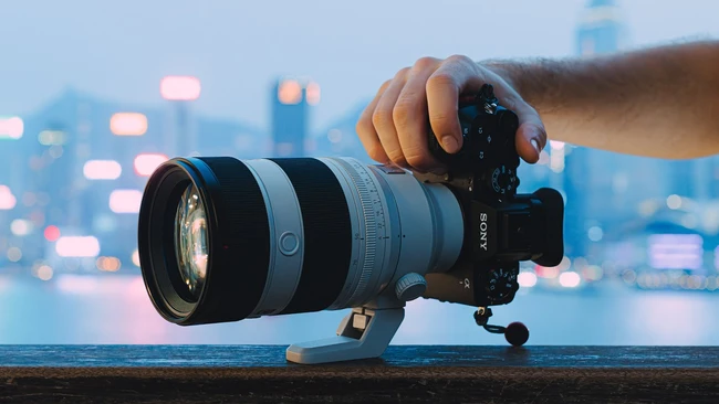 Hand holding Sony mirrorless camera with telephoto zoom lens mounted on tripod head, city skyline with bokeh lights in background