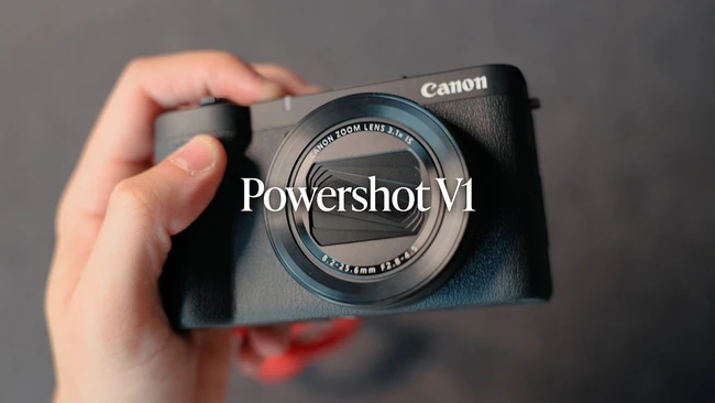 Hand holding Canon PowerShot V1 compact camera with zoom lens against gray background