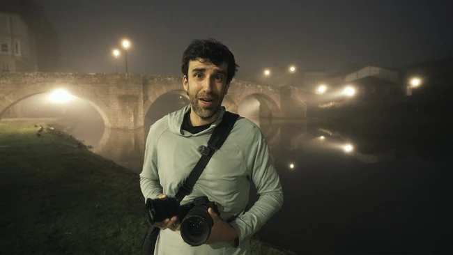 Photographer holding a DSLR camera at night in front of an illuminated stone bridge