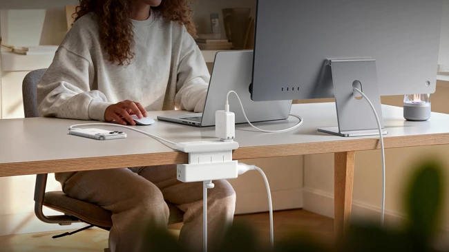 Woman at wooden desk working on laptop surrounded by white tech accessories and charging devices