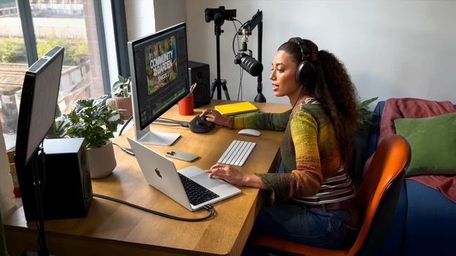 Woman in home studio wearing headphones at desk with laptop, monitor, and professional microphone setup