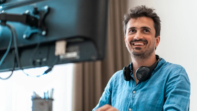 Smiling man in denim shirt at desk with monitor and headphones around neck