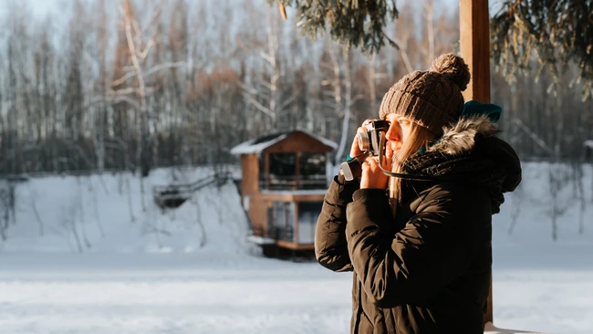 Person in winter coat and knit hat holding warm beverage outdoors in snowy landscape