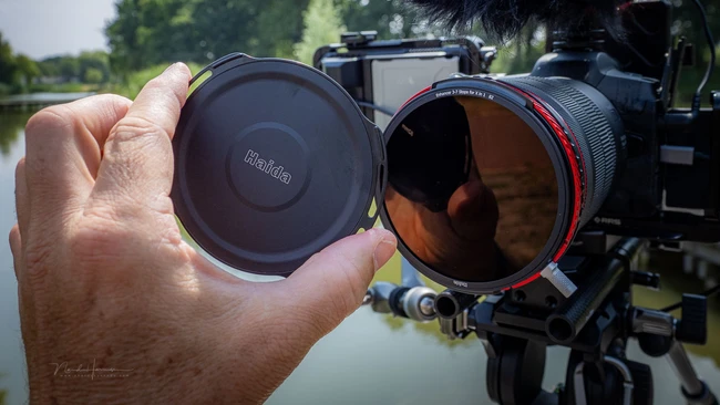 Photographer holding lens cap andND filter in front of mounted cinema camera overlooking water