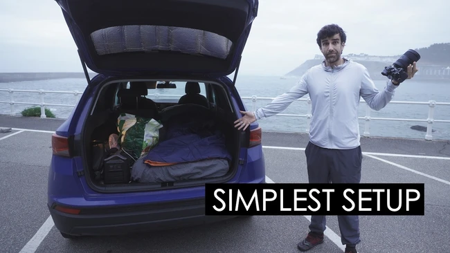 Filmmaker holding camera beside open car trunk packed with gear overlooking coastal highway
