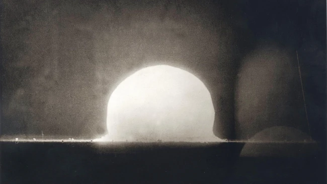 Nuclear explosion test shot showing bright hemispherical fireball rising above desert ground with shock wave and dust plume