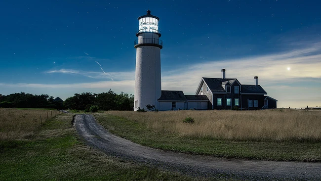 Tall white lighthouse with black band stands beside a blue Victorian keeper's house on grassy coastal terrain