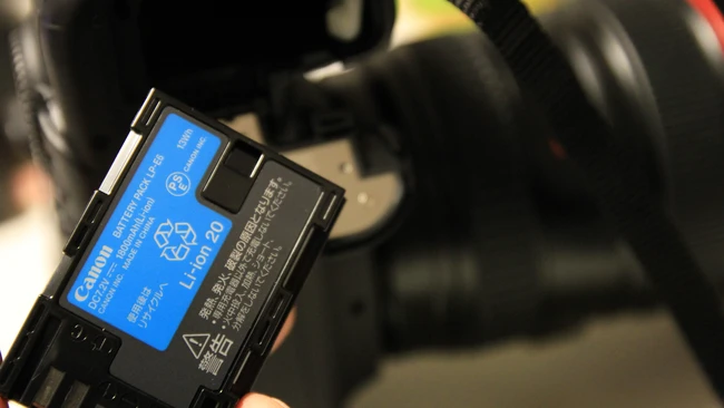 Hand holding a blue Canon LP-E6 lithium-ion battery pack in front of a blurred DSLR camera and lens.