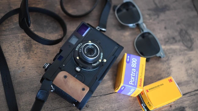 Vintage rangefinder camera with leather strap, sunglasses, and film boxes on wooden surface