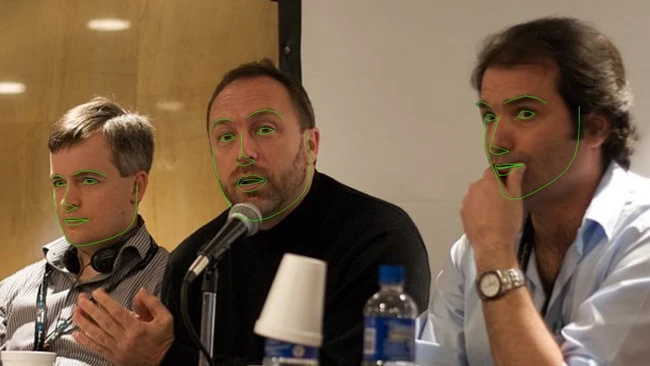 Three men on a panel, center speaker talking into a microphone, green face-tracking outlines overlaid on their faces.