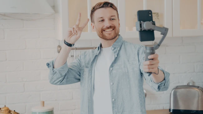 Man in light blue shirt making peace sign while holding smartphone gimbal in kitchen