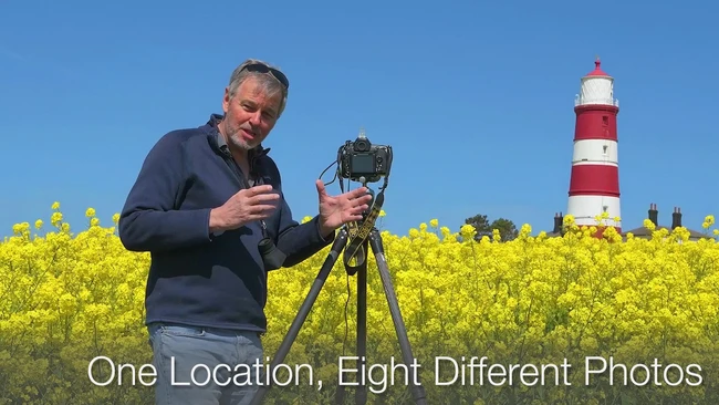 Photographer holding camera on tripod in yellow rapeseed field with red and white striped lighthouse in background